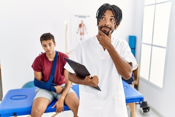 Young hispanic man working at pain recovery clinic with a man with broken arm looking confident at the camera smiling with crossed arms and hand raised on chin. thinking positive.