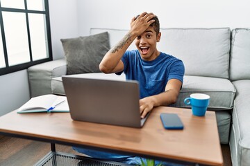 Young handsome hispanic man using laptop sitting on the floor surprised with hand on head for mistake, remember error. forgot, bad memory concept.