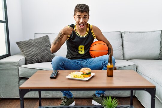 Young handsome hispanic man holding basketball ball cheering tv game pointing finger to one self smiling happy and proud