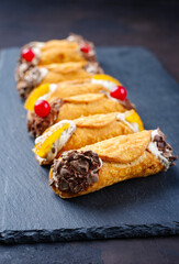 Traditional Italian Sicilian pastry dessert cannoli with creamy ricotta filling, chocolate crisps and fresh currant fruit served as close-up on a rustic black board