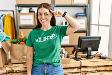 Young brunette woman wearing volunteer t shirt at donations stand smiling pointing to head with one...