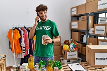 Young arab man wearing volunteer uniform talking on the smartphone holding canned food at charity center