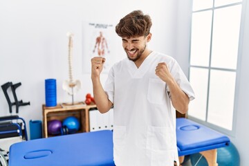 Young arab man working at pain recovery clinic very happy and excited doing winner gesture with arms raised, smiling and screaming for success. celebration concept.