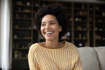 Head shot happy dreamy young African American sincere woman looking in distance, resting on cozy sofa. Inspired young pretty woman daydreaming, visualizing future, recollecting good memories indoors.