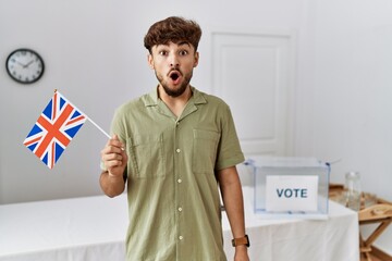 Young arab man at political campaign election holding uk flag scared and amazed with open mouth for surprise, disbelief face