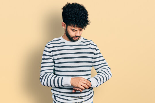Young arab man with beard wearing casual striped sweater checking the time on wrist watch, relaxed and confident