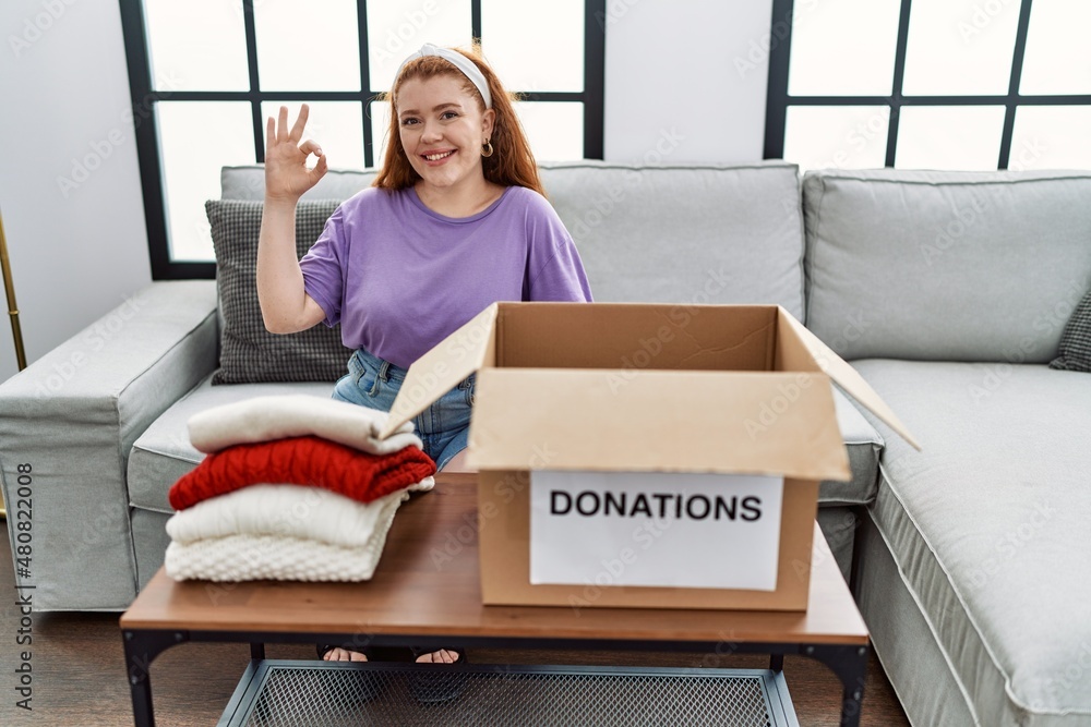 Canvas Prints Young redhead woman putting used clothes into donations box doing ok sign with fingers, smiling friendly gesturing excellent symbol