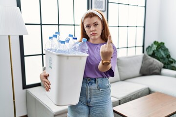 Young redhead woman holding recycling wastebasket with plastic bottles showing middle finger,...
