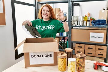 Young redhead woman wearing volunteer t shirt holding second hand sneakers doing ok sign with...