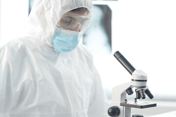 Doctor in a protective suit wearing a mask wearing blue gloves in a laboratory looks through a microscope