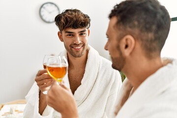 Two hispanic men couple toasting with champagne sitting on massage table at beauty center