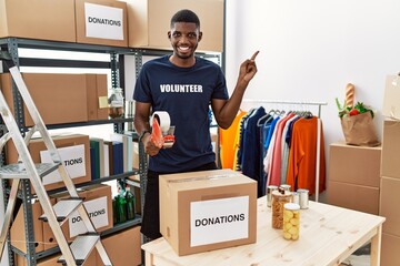 Young african american volunteer man packing donations box for charity cheerful with a smile on face pointing with hand and finger up to the side with happy and natural expression
