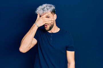 Young hispanic man with modern dyed hair wearing casual blue t shirt peeking in shock covering face and eyes with hand, looking through fingers afraid