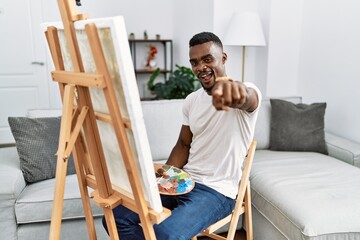 Young african man painting on canvas at home pointing to you and the camera with fingers, smiling positive and cheerful
