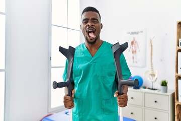 Young african physiotherapist man holding crutches sticking tongue out happy with funny expression.