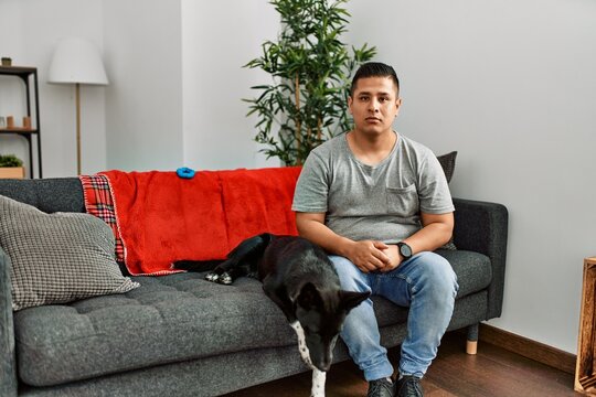 Young latin man and dog sitting on the sofa at home relaxed with serious expression on face. simple and natural looking at the camera.