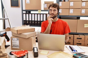 Young hispanic call center agent man working at warehouse with hand on chin thinking about question, pensive expression. smiling with thoughtful face. doubt concept.