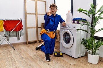 Young hispanic man working on washing machine pointing with finger to the camera and to you,...