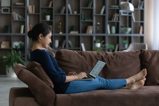 Side view relaxed happy young Indian ethnicity woman working with electronic documents on computer, editing text preparing report, writing novel, sitting on comfortable couch in living room.