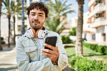 Fototapeta premium Handsome hispanic man with beard smiling happy outdoors on a sunny day wearing headphones listening to music with smartphone