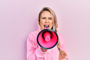 Fototapeta premium Young beautiful blonde caucasian woman shouthing and screaming through megaphone over isolated pink background