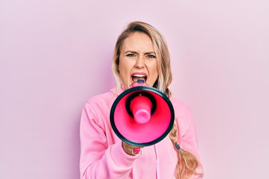 Young beautiful blonde caucasian woman shouthing and screaming through megaphone over isolated pink background