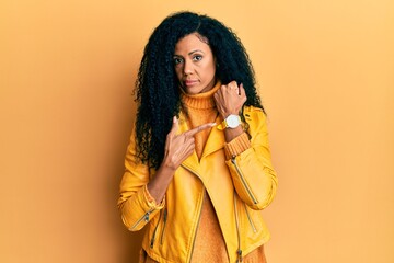 Middle age african american woman wearing wool winter sweater and leather jacket in hurry pointing to watch time, impatience, looking at the camera with relaxed expression