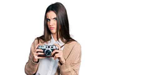 Young brunette teenager holding vintage camera clueless and confused expression. doubt concept.
