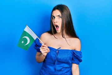 Young brunette teenager holding pakistan flag scared and amazed with open mouth for surprise, disbelief face