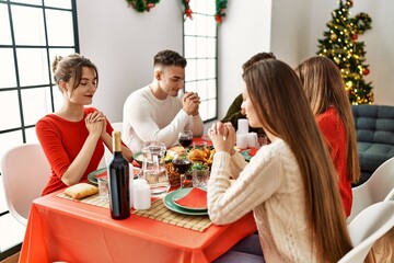 Group of young people celebrating christmas praying for food sitting on the table at home.