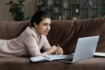 Concentrated young Indian woman wearing wired earphones looking at laptop screen, involved in educational virtual event, listening lecture webinar online, writing notes lying on couch.