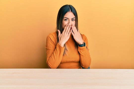 Young hispanic woman wearing casual clothes sitting on the table laughing and embarrassed giggle covering mouth with hands, gossip and scandal concept