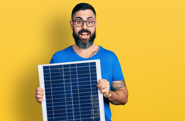 Hispanic man with beard holding photovoltaic solar panel celebrating crazy and amazed for success...