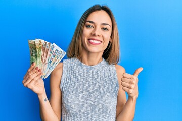 Young caucasian blonde woman holding peruvian sol banknotes smiling happy and positive, thumb up doing excellent and approval sign - obrazy, fototapety, plakaty