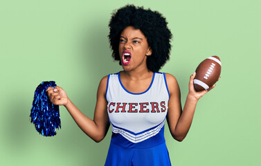 Young african american woman wearing cheerleader uniform holding pompom and football ball angry and...