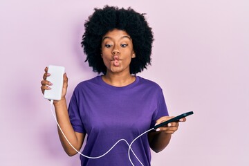 Young african american woman charging smartphone using portable battery making fish face with mouth...
