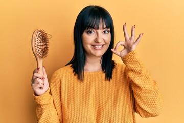 Young hispanic woman holding comb loosing hair doing ok sign with fingers, smiling friendly...