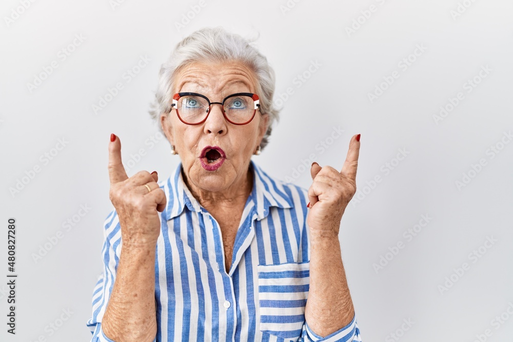 Wall mural Senior woman with grey hair standing over white background amazed and surprised looking up and pointing with fingers and raised arms.