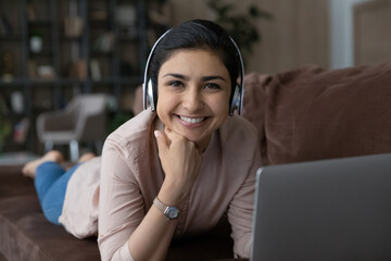 Portrait of happy young Indian woman in headphones lying on comfortable sofa with computer, enjoying spending time online watching movies, communicating with friends, listening music or lecture.