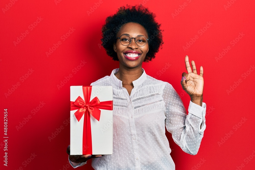 Canvas Prints Young african american woman holding gift doing ok sign with fingers, smiling friendly gesturing excellent symbol