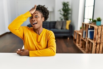 Young african american woman wearing casual clothes sitting on the table at home surprised with...