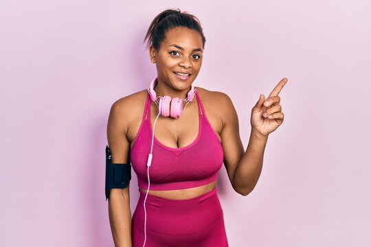 Young african american girl wearing gym clothes and using headphones with a big smile on face, pointing with hand finger to the side looking at the camera.