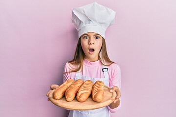 Beautiful brunette little girl wearing baker uniform holding homemade bread afraid and shocked with surprise and amazed expression, fear and excited face.
