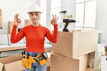 Middle age grey-haired woman wearing hardhat standing at new home relax and smiling with eyes closed doing meditation gesture with fingers. yoga concept.