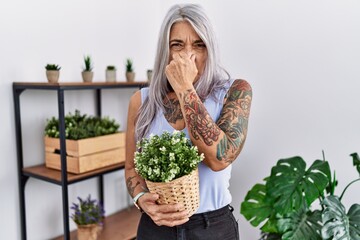 Middle age grey-haired woman holding green plant pot at home smelling something stinky and...