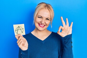 Young blonde woman holding anarchism drawing reminder doing ok sign with fingers, smiling friendly...
