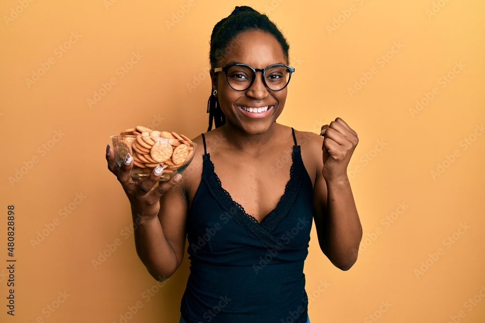 Sticker African american woman with braided hair holding salty biscuits bowl screaming proud, celebrating victory and success very excited with raised arm