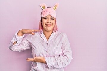 Hispanic woman with pink hair wearing sleep mask and pajama gesturing with hands showing big and large size sign, measure symbol. smiling looking at the camera. measuring concept.