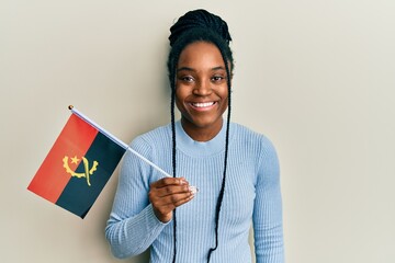 African american woman with braided hair holding angola flag looking positive and happy standing and smiling with a confident smile showing teeth