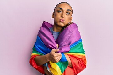 Hispanic man wearing make up and long hair wrapped in rainbow lgbtq flag puffing cheeks with funny face. mouth inflated with air, catching air.
